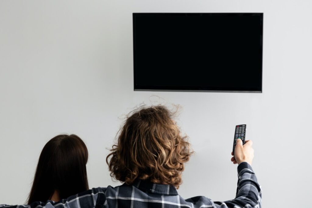 Couple Pointing Remote Control On Empty TV Screen Indoor, Back-View