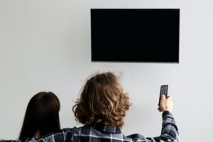 Couple Pointing Remote Control On Empty TV Screen Indoor, Back-View